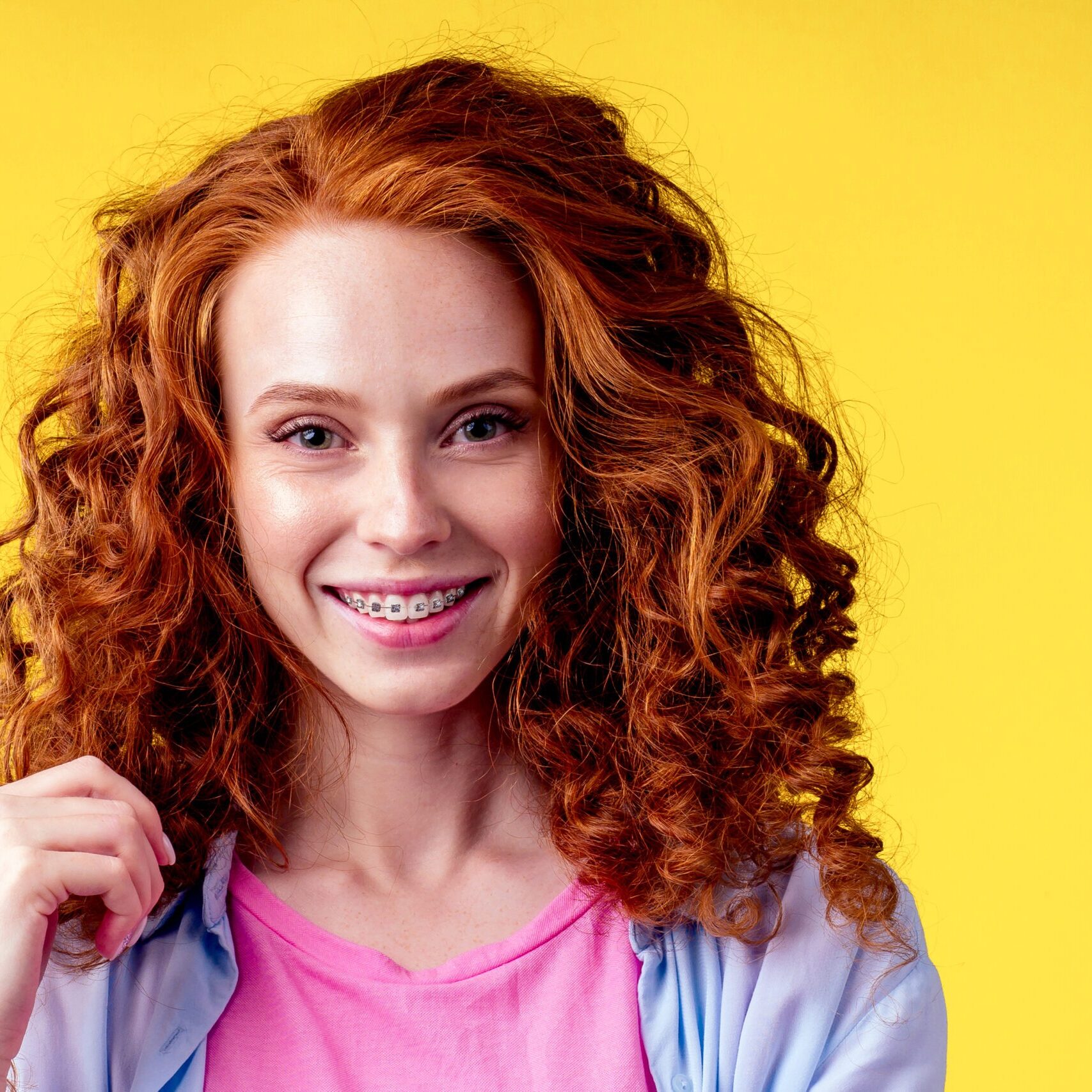 curly redhaired ginger woman with snow white smile brackets on teeth touching a new hairstyle in studio yellow background curly redhaired ginger woman with snow white smile brackets on teeth touching a new hairstyle in studio yellow background.
